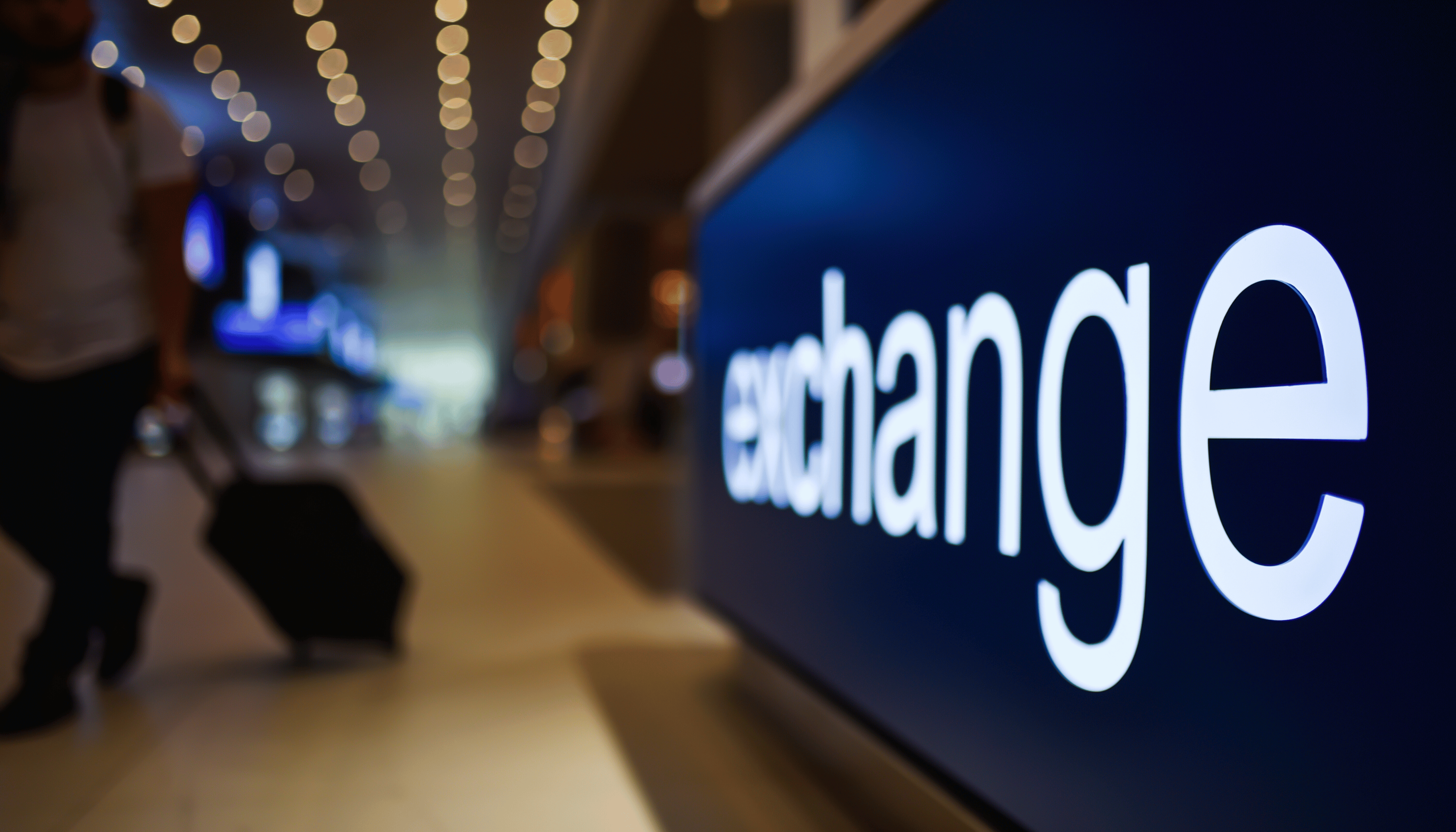 Illuminated 'exchange' sign in an airport, with a person pulling a suitcase in the background, symbolizing the currency exchange services available in travel hubs.