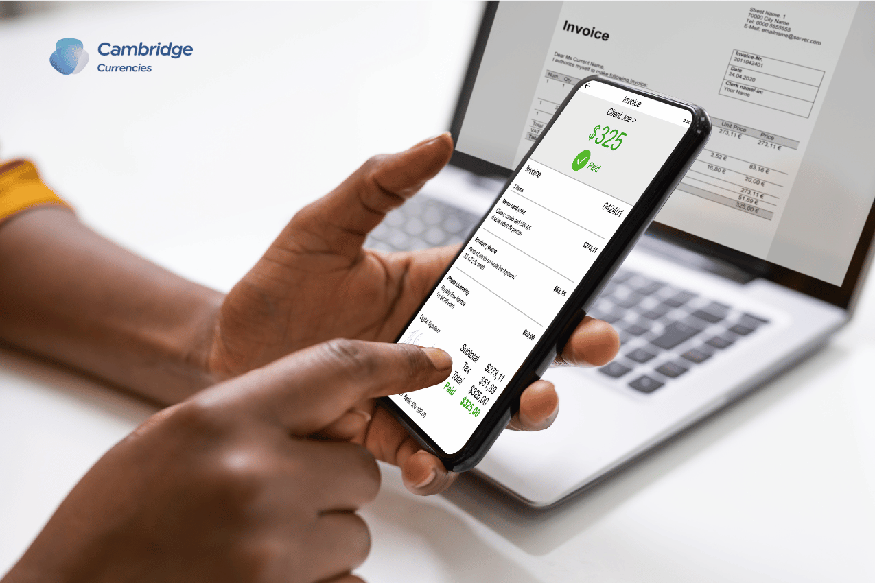 A person reviewing a digital invoice on a smartphone while sitting in front of a laptop displaying another invoice. Cambridge Currencies logo is visible, representing international business payment solutions.