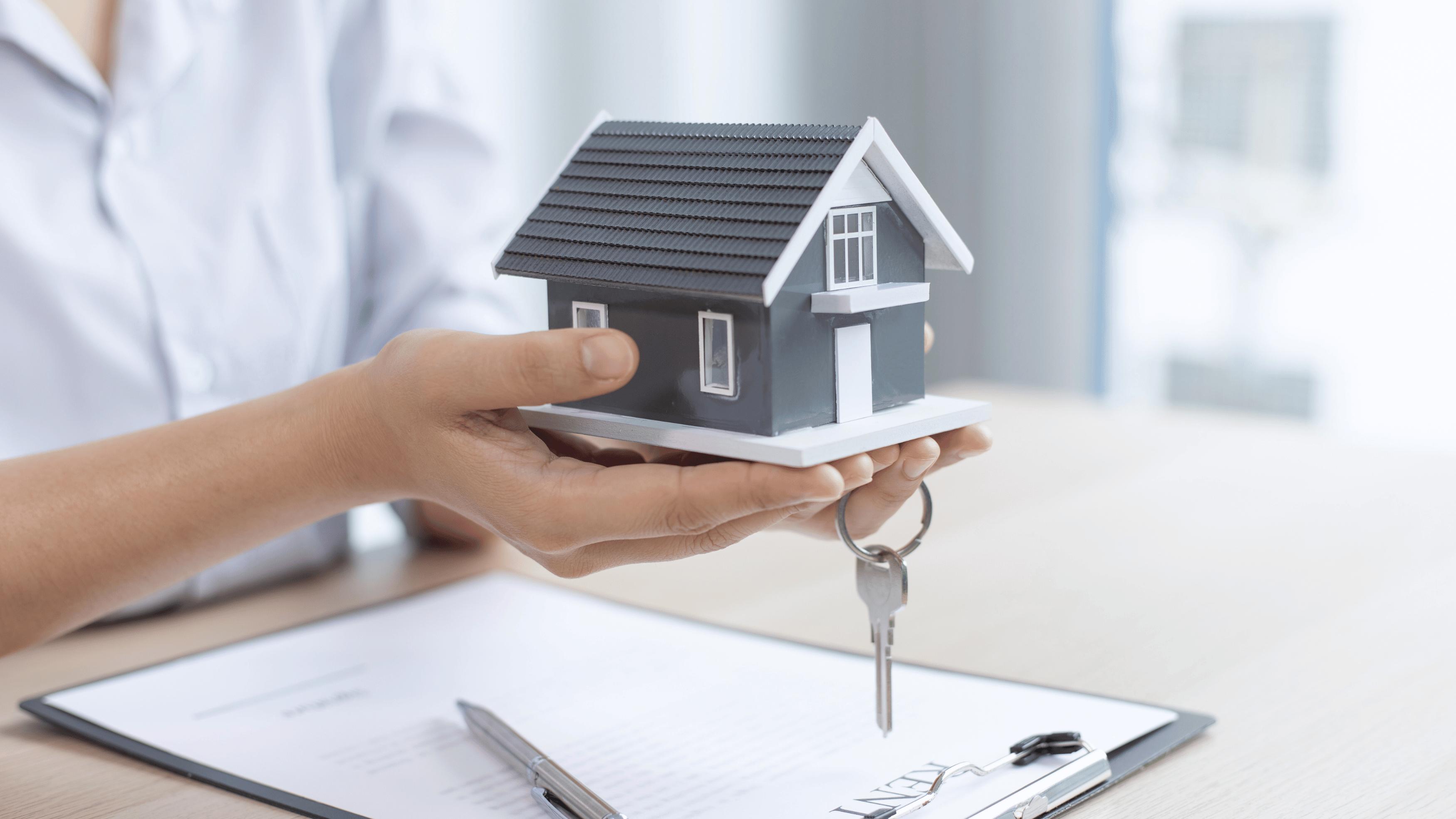 Person holding a miniature house model with keys, symbolizing property sale or investment alongside a signed contract.