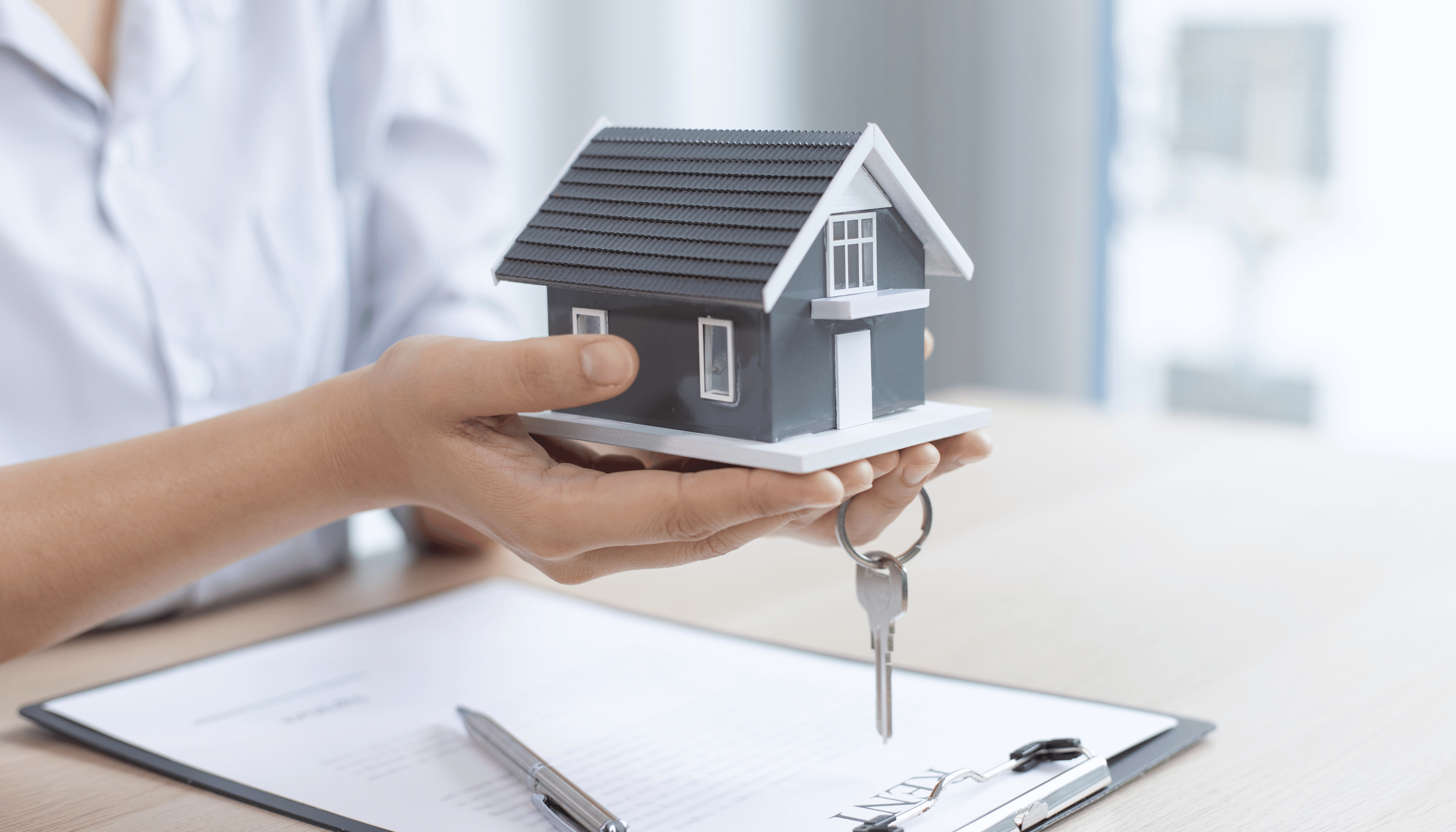 Person holding a miniature house model with keys, symbolizing property sale or investment alongside a signed contract.